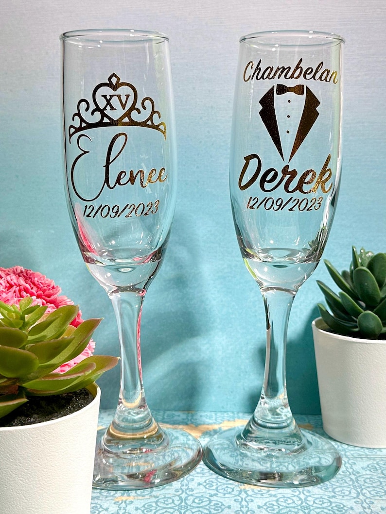 May include: Two clear champagne flutes with gold lettering. One flute has a crown design with the name "Elenee" and the date "12/09/2023". The other flute has a tuxedo design with the name "Derek" and the date "12/09/2023".
