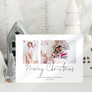 Op de afbeelding: Een witte kerstkaart met twee familiefoto's en de woorden "Merry Christmas" in sierlijke letters. De kaart bevat ook de tekst "Lots of love from the Thompsons" en het jaar "December 2025". Decoratieve witte keramische huisjes en bomen omringen de kaart.