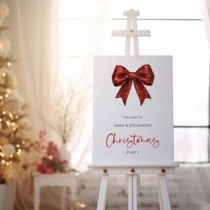 Puede incluir: Un letrero blanco sobre un caballete de madera con un lazo rojo brillante. El texto "Welcome to Emily & Jonathon's Christmas Party" está escrito en rojo. Un árbol de Navidad es visible en el fondo.