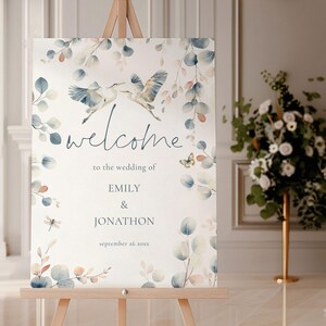 May include: A watercolor wedding welcome sign with a floral design featuring blue and beige leaves, two blue herons flying, a dragonfly, and a butterfly. The text reads "welcome to the wedding of Emily & Jonathon September 26 20XX".