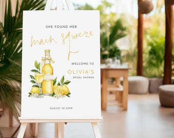 May include: A white easel sign with gold lettering and a watercolor illustration of lemons and a bottle of limoncello. The sign reads "She found her main squeeze" and welcomes guests to Olivia's Bridal Shower, with the date August 28 20XX.
