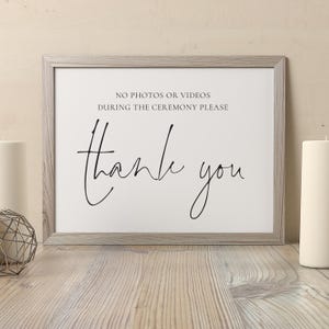Könnte beinhalten: Ein gerahmtes Schild mit den Worten "Thank you" in eleganter Schrift. Das Schild enthält auch den Text "NO PHOTOS OR VIDEOS DURING THE CEREMONY PLEASE". Der Rahmen ist hellgrau und der Hintergrund ist weiß.