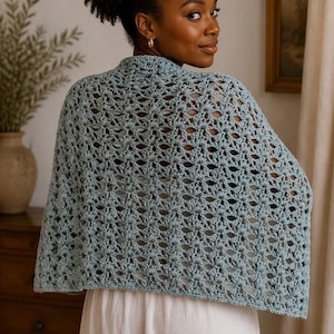 May include: A light blue crochet shawl featuring an intricate, repeating openwork pattern. The garment is draped over the shoulders, showcasing a textured, airy design with a scalloped edge finish, crafted from soft yarn in a muted dusty blue hue.