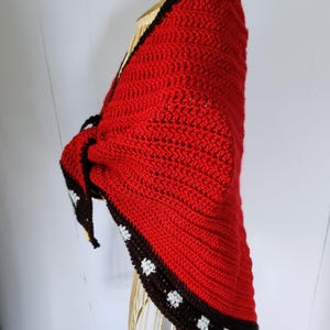 May include: A triangular crochet shawl featuring a vibrant red main body with a textured stitch pattern. The edges are finished with a wide dark brown border, accented by small white floral motifs and a thin yellow stripe along the bottom hem.