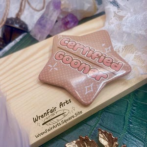 Könnte beinhalten: Ein sternförmiger Anstecker mit dem Text "Certified Gooner" in Hellrosa, der auf einer Holzoberfläche liegt. Ein goldfarbener Blattanhänger und Kristalle sind ebenfalls zu sehen. Das Bild ist mit einer Feder und Spitze gestaltet.