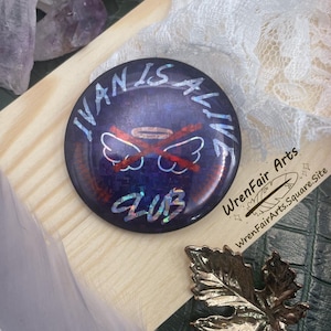 May include: A round, dark blue button with the text "IVAN IS ALIVE CLUB" in white script. The button features a red cross over a halo and wings design. The button is on a wooden surface with crystals, a feather, and a gold leaf pendant.