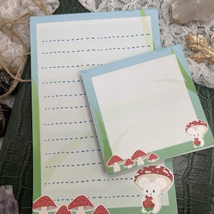 May include: Two notepads with a whimsical design. The larger notepad has lined paper with a light blue border. The smaller notepad has a white background with a green border. Both notepads feature illustrations of red mushrooms and a cute mouse holding a strawberry.