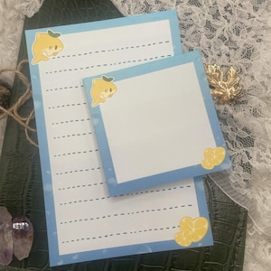 May include: Two notepads with a light blue border and a lemon shark illustration. One notepad is lined, and the other is blank. The notepads are decorated with yellow lemon slice stickers. The notepads are on a dark green surface.