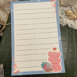 May include: A light blue notepad with a border of white polka dots and a pink ribbon design. The notepad features a stack of pink macarons topped with a cherry and a chocolate-covered strawberry illustration.