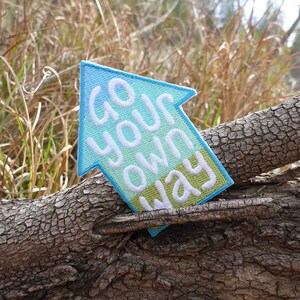 May include: A blue, green, and white embroidered arrow patch with the text "Go your own way". The patch is sitting on a tree branch.