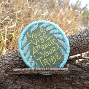 Puede incluir: Un parche redondo de tela verde con costuras azules alrededor del borde. El parche tiene el texto "Your Vibe Attracts Your Tribe" bordado en hilo amarillo.