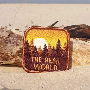 May include: A brown embroidered patch with a sunset scene and a forest silhouette. The text "THE REAL WORLD" is printed in orange on the patch.
