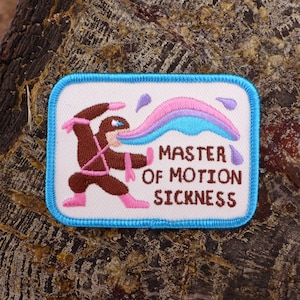 May include: A white embroidered patch featuring a brown cartoon figure in a pink and brown outfit. The figure is blowing a rainbow-coloured stream of air. The text "MASTER OF MOTION SICKNESS" is below the figure.