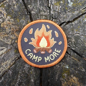 Puede incluir: Un parche redondo bordado de color gris oscuro con un borde naranja. El parche presenta una fogata con llamas naranja y amarillas y el texto "Camp More" en amarillo.