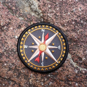 Puede incluir: Un parche de brújula bordado en negro y dorado con una estrella blanca y una flecha roja. La brújula está sobre una superficie rugosa, marrón y texturizada.