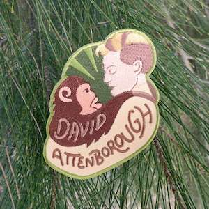 Op de afbeelding: Een bruin en groen geborduurde patch met een cartoon afbeelding van een man en een aap. De tekst "DAVID ATTENBOROUGH" is in bruin geborduurd op de patch.