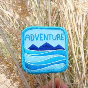 May include: A blue and white embroidered patch with the word "ADVENTURE" in white text and a blue mountain range and blue waves design.