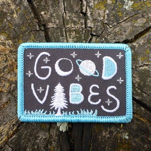 May include: A gray embroidered patch with a blue border. The patch features the text "Good Vibes" in white embroidery with a stylized tree, moon, and planet.