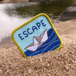 Op de afbeelding: Een blauwe en gele geborduurde patch met het woord "ESCAPE" in witte letters. Een wit papieren bootje vaart op blauw water met witte golven.