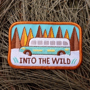 May include: An embroidered patch with a blue and white vintage bus driving through a forest of brown and orange trees. The text "INTO THE WILD" is stitched in brown thread below the bus.