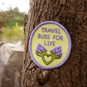 Puede incluir: Un parche bordado amarillo y morado con el texto "Travel Buds for Life" y un corazón verde con capullos de flores moradas a cada lado.