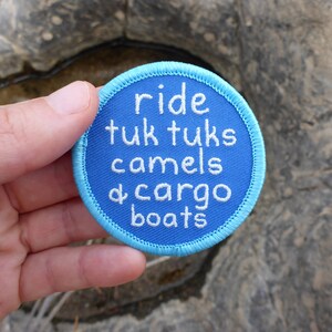 May include: A blue and light blue round embroidered patch with white text that reads "ride tuk tuks camels & cargo boats".