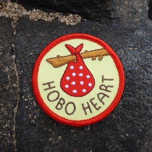 May include: An embroidered patch featuring a red and white hobo bag hanging from a stick. The text "HOBO HEART" is stitched in brown around the edge of the patch.