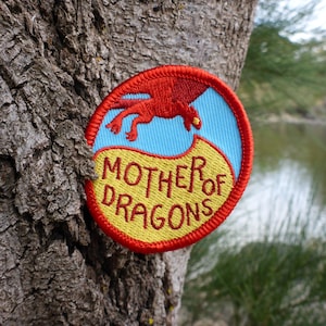 Puede incluir: Un parche bordado rojo, amarillo y azul con un dragón rojo y el texto "Mother of Dragons".