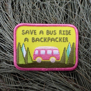Op de afbeelding: Een geborduurde patch in geel en roze met de tekst "Save a bus ride a backpacker" en een roze cartoonbusje met groene bomen aan beide kanten.