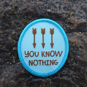 Op de afbeelding: Een blauwe en bruine geborduurde patch met drie bruine pijlen die naar beneden wijzen en de tekst "YOU KNOW NOTHING" in bruin.