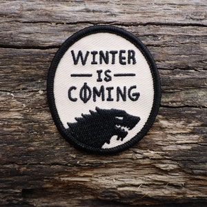 May include: A white circular patch with black embroidery. The text "Winter is Coming" is stitched in black thread. A black direwolf silhouette is embroidered at the bottom of the patch.