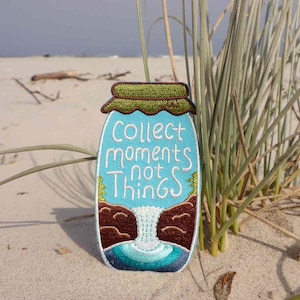 May include: A blue embroidered patch shaped like a jar with a green lid. The jar has a waterfall inside and the text "Collect moments not things" is embroidered on the front.