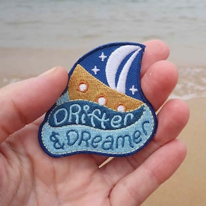 May include: A blue and yellow embroidered patch with a sailboat and stars. The text on the patch reads "Drifter & Dreamer".