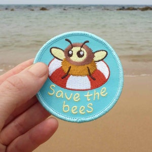 Puede incluir: Un parche bordado azul y blanco con una abeja de dibujos animados que lleva un salvavidas rojo y blanco. El parche dice "Save the bees" en texto blanco.