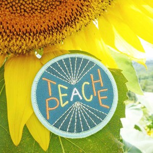 Könnte beinhalten: Ein runder blauer und weißer Patch mit dem Text "TEACH PEACE" in orangefarbenen und rosafarbenen Buchstaben. Der Patch befindet sich auf einem grünen Blatt mit einer gelben Sonnenblume im Hintergrund.