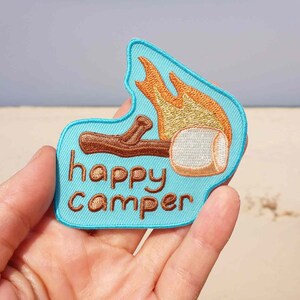 May include: A blue embroidered patch with the text "happy camper" in brown thread. The patch features a brown stick with a white marshmallow on top, with orange flames behind it.