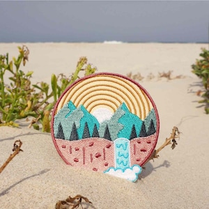 May include: A round embroidered patch with a mountain scene. The patch features a sunset with a rainbow, mountains, trees, and a waterfall. The patch is on a sandy beach.