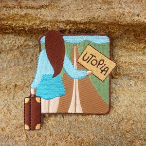 May include: Embroidered patch depicting a woman with brown hair, wearing a blue shirt and skirt, holding a sign that reads "Utopia". She is walking towards a road with a suitcase in her hand. The patch is set against a brown background.