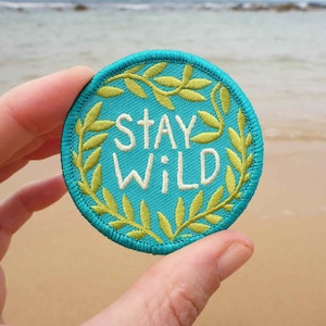 May include: A turquoise blue embroidered patch with a green leafy border. The patch says "Stay Wild" in white embroidery.