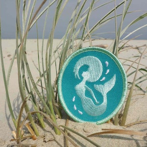 Könnte beinhalten: Ein türkisfarbener gestickter Aufnäher mit einem weißen Meerjungfrauen-Motiv. Die Meerjungfrau schwimmt mit ihrem Schwanz nach oben und ihren Haaren, die hinter ihr herwehen. Der Aufnäher ist von weißem Sand und grünem Gras umgeben.
