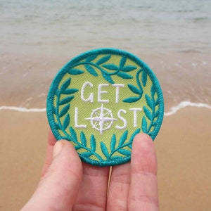 Pode incluir: Um remendo redondo bordado de cor verde e amarela com uma bússola e o texto "GET LOST" em branco.