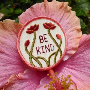 Puede incluir: Un parche de tela blanca con tallos bordados en verde y flores bordadas en rojo. El parche tiene un borde rojo y las palabras "BE KIND" bordadas en hilo dorado.