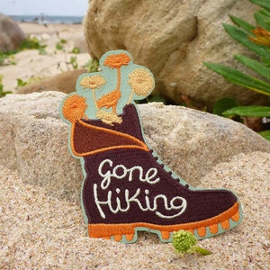 Op de afbeelding: Een bruine wandelschoen met oranje veters en een groene, bladerrijke plant die uit de bovenkant groeit. De schoen heeft de tekst "Gone Hiking" geborduurd op de zijkant in witte draad.