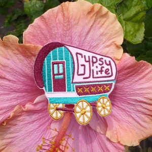 Op de afbeelding: Gestikt patch met een turquoise en witte wagen met een bordeauxrood dak en een raam. De wagen heeft wielen met gouden velgen en de tekst "Gypsy Life" is geborduurd op de zijkant.