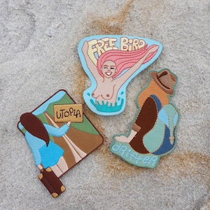 May include: Three embroidered iron-on patches. One patch features a woman with pink hair and the text "FREE BIRD". Another patch features a woman with brown hair holding a sign that says "UTOPIA". The third patch features a woman with brown hair and the text "DRIFTER".