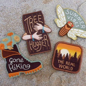 Op de afbeelding: Vier geborduurde patches met natuurthema's. De patches tonen een bruine laars met de tekst "Gone Hiking", een bruine boomstam met de tekst "Tree Hugger", een groene cicada met de tekst "Summer Vibes" en een zonsondergangsscene met de tekst "The Real World".