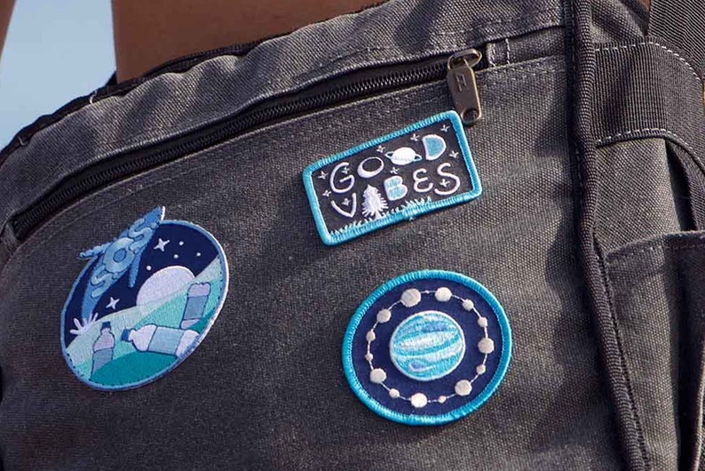 May include: A black canvas bag with three embroidered patches. The patches are blue and white and feature a moon, stars, a planet, and the words "Good Vibes".