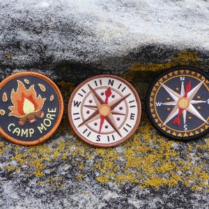 May include: Three embroidered patches on a gray textured surface. The patches are circular and feature a campfire with the text "Camp More", a compass with the directions North, South, East, and West, and a compass with a star in the center.
