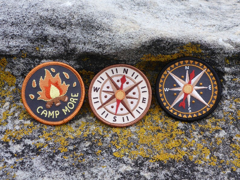 May include: Three embroidered patches on a gray textured surface. The patches are circular and feature a campfire with the text "Camp More", a compass with the directions North, South, East, and West, and a compass with a star in the center.