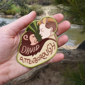 K&ouml;nnte beinhalten: Ein gestickter Aufn&auml;her in Gr&uuml;n, Braun und Beige mit einem Cartoon-Bild eines Mannes mit einem Affen. Der Text "David Attenborough" ist auf den Aufn&auml;her gestickt.